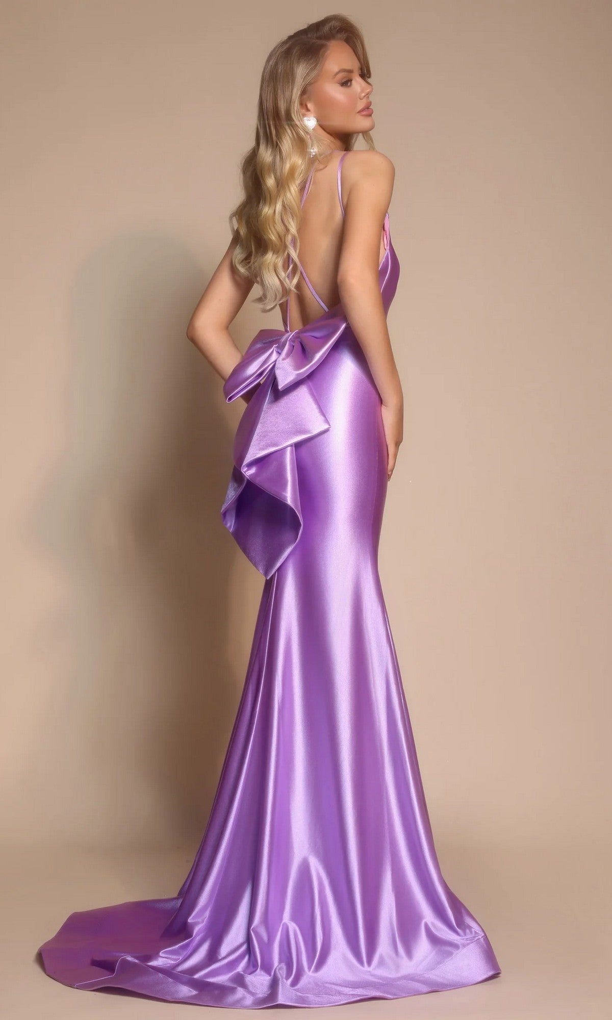 A woman stands facing away against a beige background, wearing the Portia and Scarlett XO095 Long Formal Dress—a shiny, floor-length lavender gown with a statement bow on the open back.